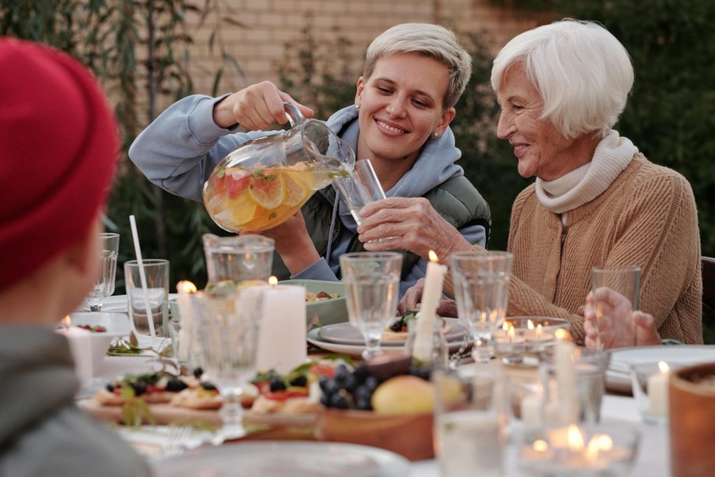 Eine gut gelaunte Frau schenkt einer älteren Dame ein Getränk ein, während sie mit ihrer Familie auf der Terrasse zu Abend isst.